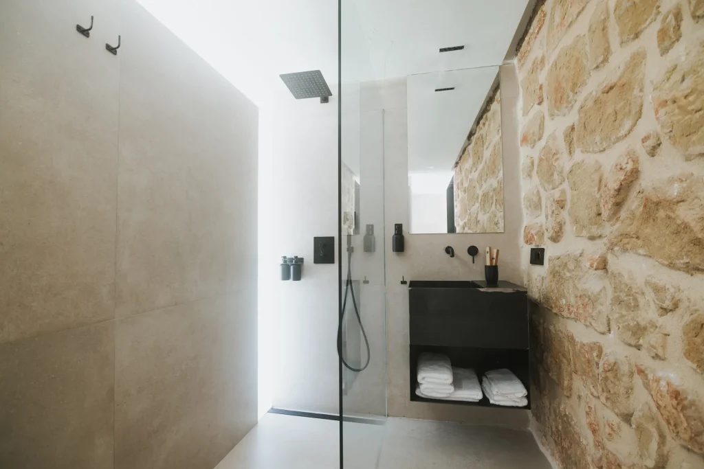 Baño del apartamento 5 con lavabo, espejo y ducha junto a un muro de piedra.